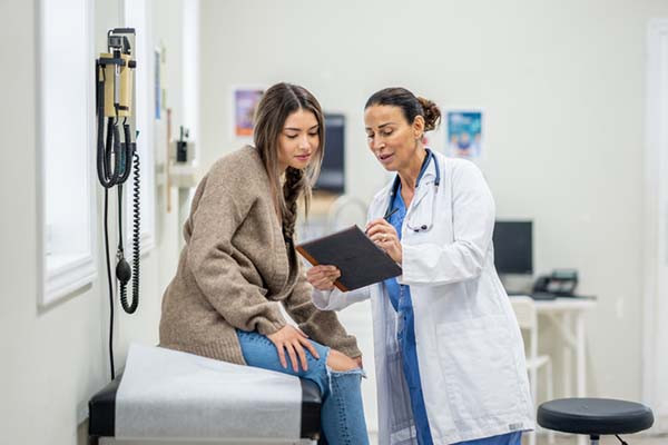 photo of a female doctor conferring with a teen female patient in an exam room, the younger woman is sitting on the exam table and the doctor is holding a tablet and showing it to her